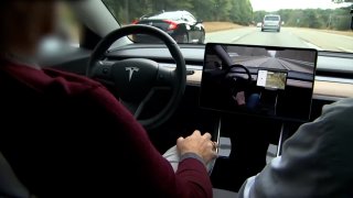 a person sits in the driver seat of a Tesla vehicle, The steering wheel, center monitor and part of the windshield are visible.