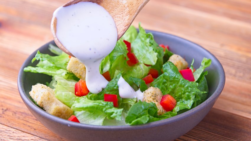 A close up of of pouring ranch dressing from a wooden spoon on a salad with peppers and croutons.